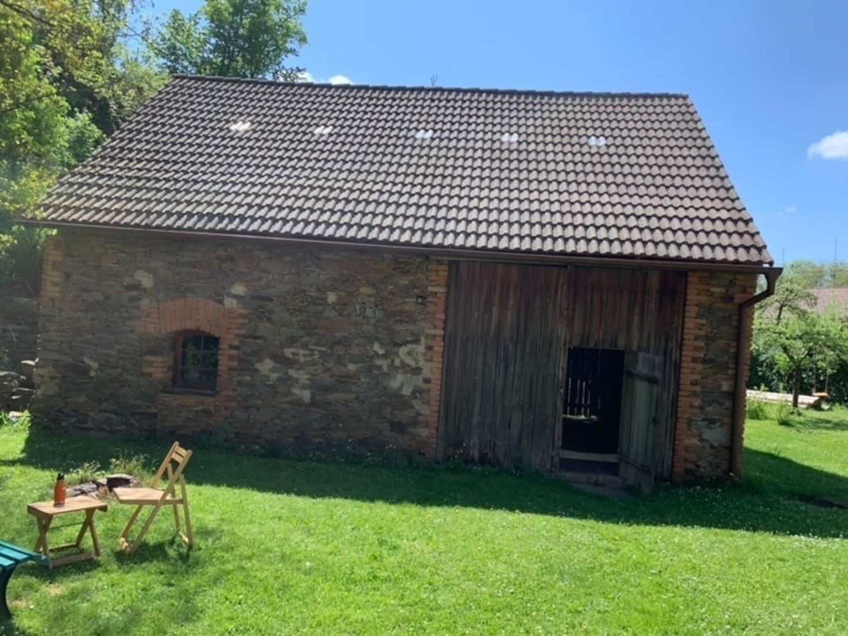 Kamenná stodola s dřevěnými vraty, před ní stolek a židle v trávě