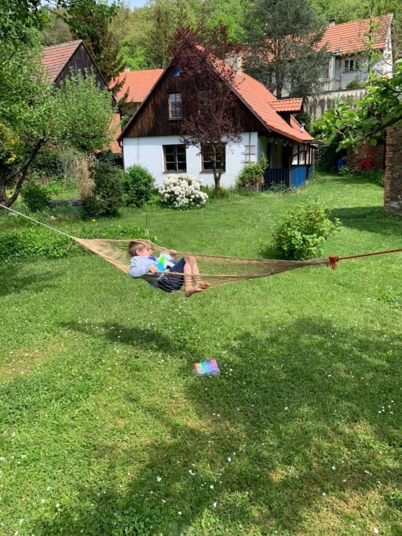 Zahrada s houpací sítí natažená mezi stromy, v pozadí bílá chaloupka s červenou střechou